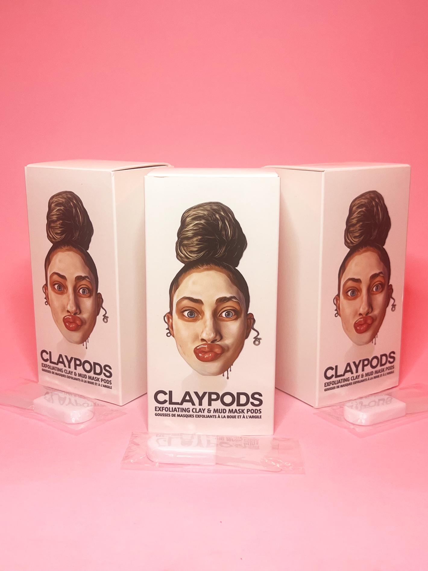 3 boxes of clay pods with mud mask brushes on a pink background